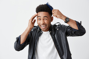 Close up isolated portrait of young funny dark-skinned attractive men with curly hair in fashionable outfit combing hair, getting ready for a walk, making silly faces. White background