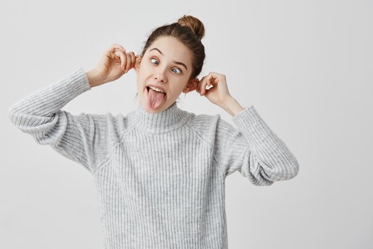 Funny emotions of hilarious female touching her ears and sticking tongue out. Caucasian woman with brown hair in bun pulling face grimacing in studio. Joy, amusement concept 