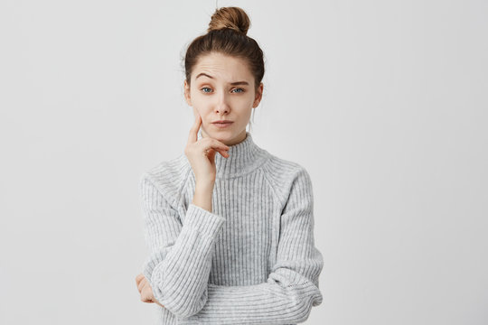 Melancholy girl 20s standing with brooding expression touching her cheek with finger. Frowning female copywriter looking on camera with one eyebrow raised being reflective. Puzzlement