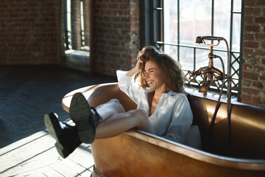 Lifestyle shot of cheerful emotional glamourous young woman with voluminous hair in black boots and white shirt sitting in brass bathtub with legs sticking out, laughing happily and having fun
