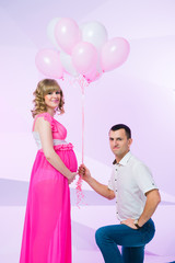 Beautiful and happy couple waiting for their daughter, standing in the studio with balloons on a pink background.