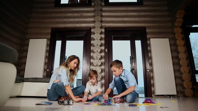 Happy mother with two children sitting on the floor of his country house spending time together collecting children's designer. Living room with panoramic Windows.
