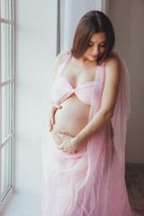 Portrait of tender pregnant beauty young woman standing in front of the window and touching her belly at home. Loft minimalistic interior