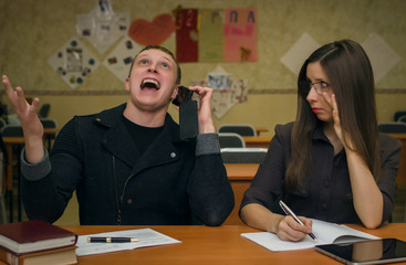 School change. Bad behavior in school concept. Two students sit at the school table in the classroom, talk on the mobile phone, fool around and prevent the teacher from leading the lesson