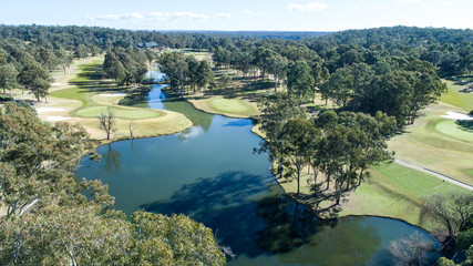Aerial view of golf course fairways and green with flag, bunkers and dam water hazard surrounded by trees in background