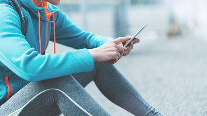 Athlete young girl sitting on the street with mobile phone, water-backpack, headphones. City workout with smart gadgets and sport applications. Smiling. Sport tight clothes.