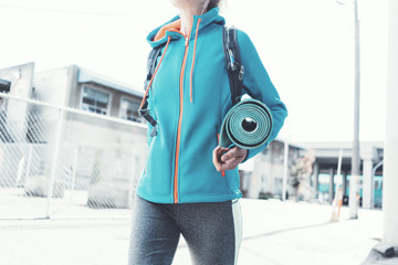 Obraz premium Athlete beautiful girl with backpack, headphones, yoga mat going to sport club on the city street. Sport tight clothes. Blurry background.