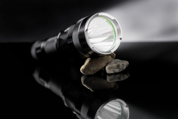 Black flashlight with lights open on stones mirroring on black background.