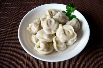 Home / purchased dumplings in a broth with greens. Dumplings are laid out in a deep white bowl. The bowl is on the stand. Day light. Dumplings are decorated with parsley leaves.