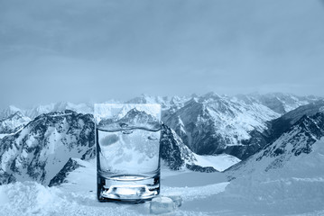 Naklejka na ściany i meble Glass with water and ice cubes on the background of a snowy mountain ridge, cold toning.