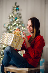 Obraz na płótnie Canvas Young smiling woman in sweater holding a gift box celebrating winter holidays in decorated home interior with Christmas tree. Happy New Year concept