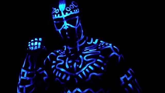 A black man in luminous patterns on a muscular body raises his hand and shows a palm with a mysterious sign and smiles, bit slow motion