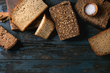 Variety loaves of sliced homemade rye bread whole grain and seeds on wooden cutting board with bowl of salt over old dark wooden background. Top view, copy space. Healthy eating