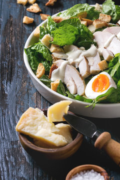 Classic Caesar salad with grilled chicken breast and half of egg in white ceramic plate. Served with ingredients above over old dark blue wooden background. Close up. Rustic style
