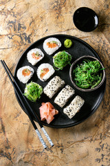 Homemade sushi rolls set with salmon, sesame seeds serving in black plate with pink pickled ginger, soy sauce, wasabi, seaweed salad, chopsticks over brown stone background. Top view, space