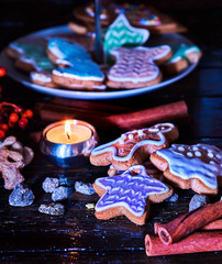 Candle light table with Christmas gingerbread cookies and cinnamon stick and star sweets are on decoration tiered cake stand wooden table and burning candles. Gingerbread trees lay on table.