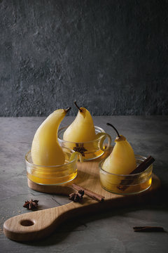 Traditional dessert poached pears in white wine served in glass bowls with syrup and lemon zest on wooden serving board over gray texture table.