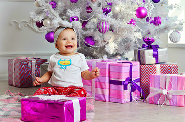 Fototapeta premium Little happy baby girl sitting on floor near white New Year tree with Christmas gifts