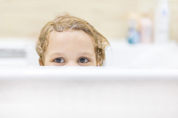 Little cute caucasian girl with blond wet hair having fun and peaking out of bath side with tricky sight