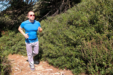 Fit runner man jogging for fitness running at running trail in beautiful landscape nature outdoors. Handsome young sportsmen wearing sportswear and running and exercises in forest. Healthy lifestyle.