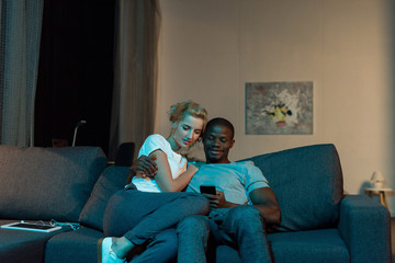 multicultural young couple using smartphone together while sitting on sofa at home