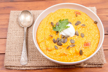 Pumpkin and carrot soup decorated with cream, parsley and pumpkin seeds on a wooden background