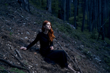 Naklejka na ściany i meble Young beautiful woman in a black long dress is sitting in a dark forest