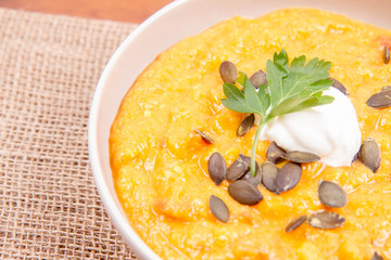 Pumpkin and carrot soup decorated with cream, parsley and pumpkin seeds on a wooden background