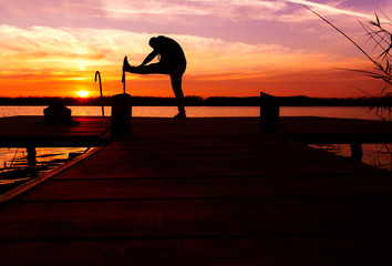 Silhouette of man stretching his muscles on a wooden dock at sunset.