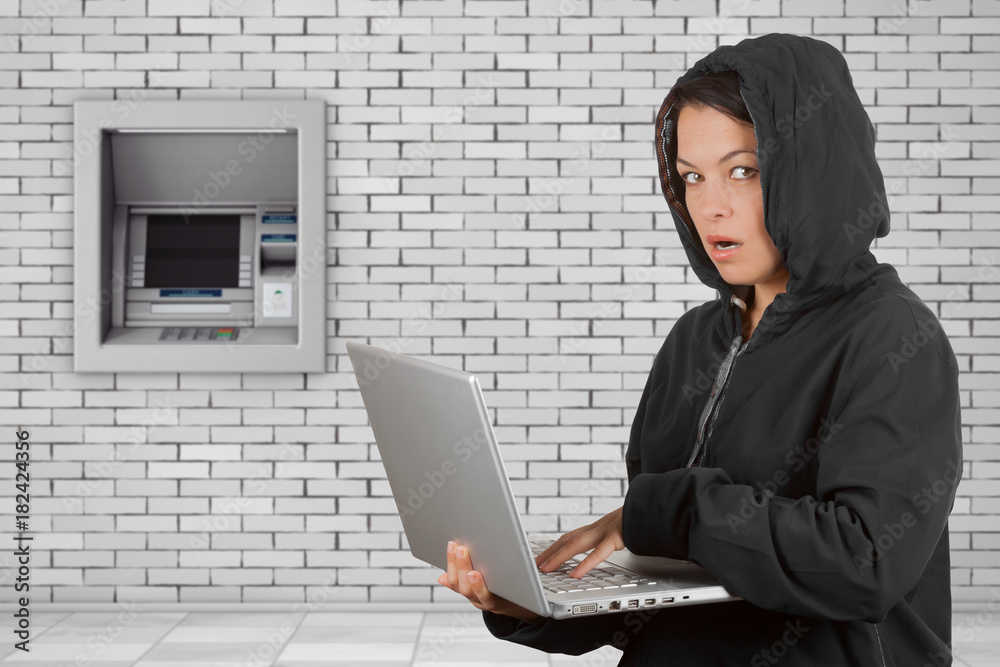 Poster criminal woman hacker wearing hood on using a laptop in front of build in bank cash atm machine.
