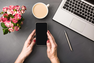 Female hands with smartphone, flowers, cup of coffee and laptop on gray table. Top view.