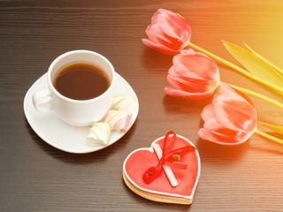 Mug of coffee, marshmallows and heartshaped gingerbread, three pink tulips. Black background.