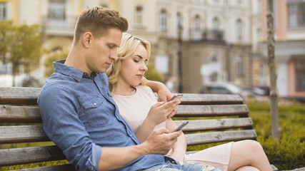 Boyfriend and girlfriend sitting on bench, engrossed in phones, networking