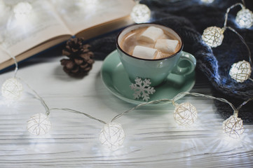 Obraz na płótnie Canvas Cozy winter home. Cup of cocoa with marshmallows, warm knitted sweater, open book, Christmas garland on a white wooden table. Atmosphere of a pleasant evening for reading.