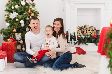 Obraz na płótnie Canvas Happy young cheerful parents with cute little son. Child boy sitting in light room at home with decorated New Year tree and gift boxes. Christmas good mood. Family, love and holiday 2018 concept.