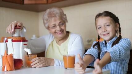 Grandmother with granddaughter are preparing fresh juice from apples ad carrots - Powered by Adobe