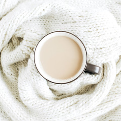 Coffee mug with milk on white wool scarf. Flat lay, top view.