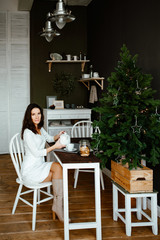 The young woman the brunette sits in kitchen with green walls in the apartment and has breakfast. On a table a cup of tea and cookies. At a background the Christmas fir-tree. 