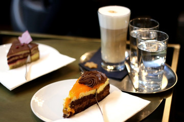 Slice of orange and chocolate cake on a coffee table. Glasses of water and coffee in the background. Selective focus. 