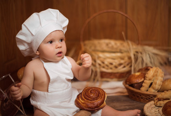 Happy little child in the cook costume at the kitchen sitting on the table among baskets with tasty rolls, buns and bread. Copy space