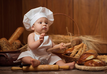 little boy in a cook costume among a baking bread rolls of flour confectionery on a brown wooden floor sits on a wooden background