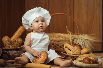 little boy in a cook costume among a baking bread rolls of flour confectionery on a brown wooden floor sits on a wooden background