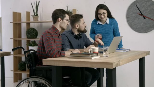 Young creative hipsters brainstorming on meeting at office and using laptop to note ideas. Disabled office worker in wheelchair and diverse colleagues discussing startup business in modern office.