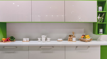 Fototapeta na wymiar Fragment of the kitchen in a modern style with dishes, fruits and vegetables on a white table top. Glaringly glossy facades and bright green furniture details.