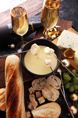 Gourmet Swiss fondue dinner on a winter evening with assorted cheeses on a board alongside a heated pot of cheese fondue with two forks dipping bread and white wine or champagne