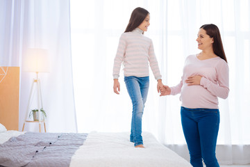 Want to grow up.  Nice girl standing on the bed while attractive pregnant woman  holding her hand and placing a hand on her own tummy