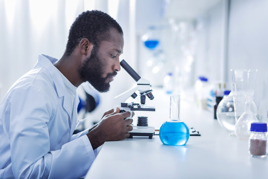 Professional laboratory. Smart handsome male scientist sitting at the table and looking into the microscope while working in the laboratory
