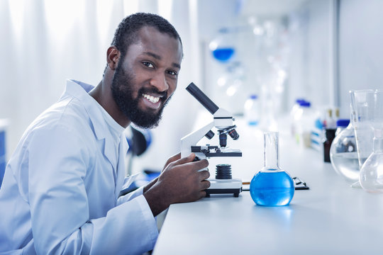 Positive mood. Cheerful nice handsome scientist sitting in front of the microscope and looking at you while enjoying his job