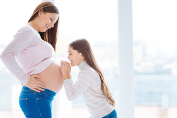 Sisters voice.  Pretty little happy girl speaking to baby in pregnant moms belly while both standing in profile and posing on the white background 