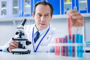 Chemical laboratory. Serious nice smart man sitting at the table and taking a test tube from the rack while doing chemical analysis